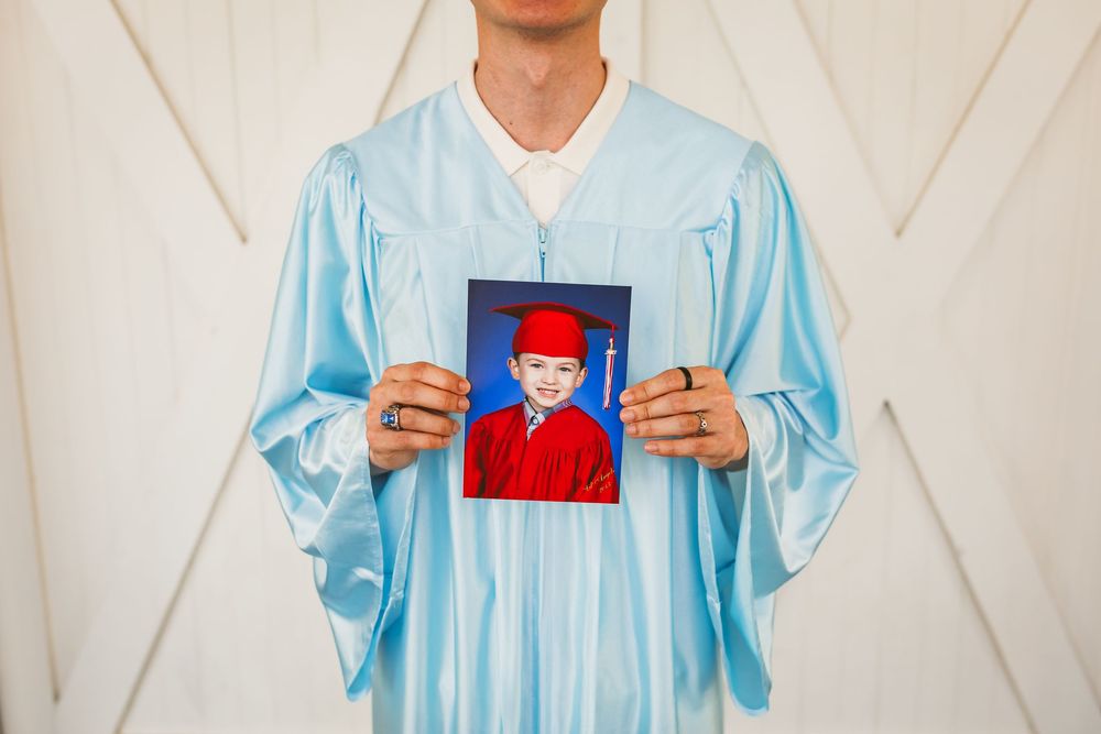 Graduate in blue cap and gown holding childhood graduation photo of child in red cap and gown.