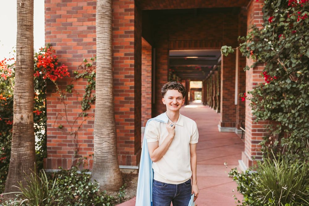 Young man in white polo shirt holding jacket, smiling in a brick archway corridor with red flowers.