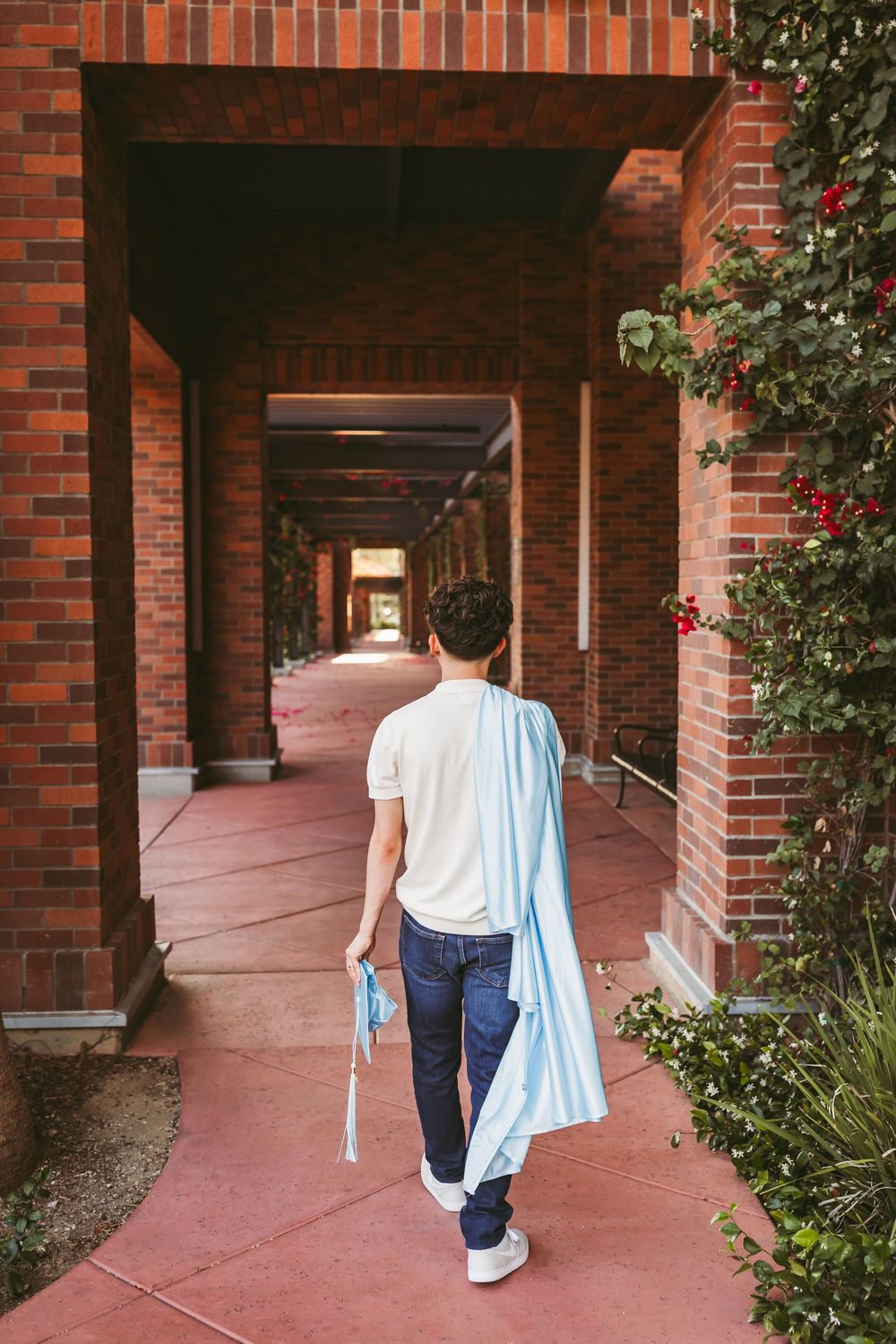 Graduate walking through brick archway corridor holding cap and gown, viewed from behind.