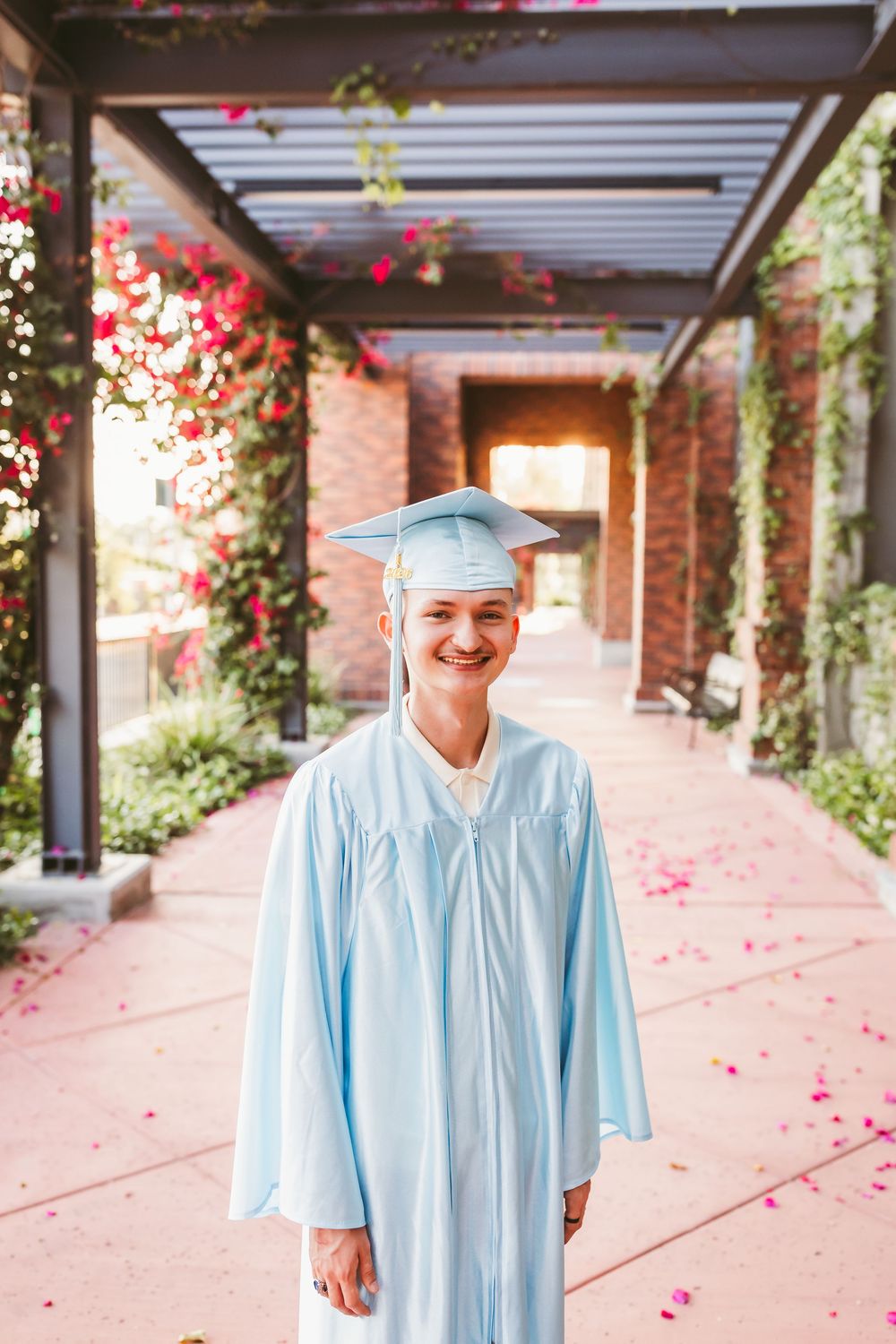 Smiling graduate in light blue cap and gown standing in a flower-lined walkway on a sunny day.