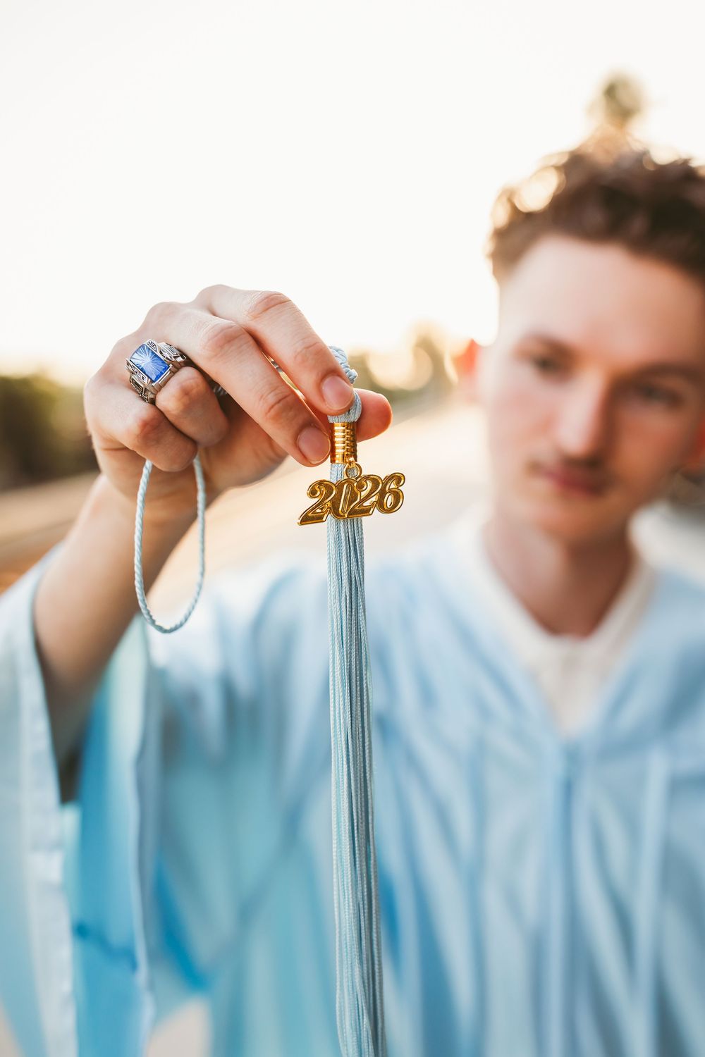 Young man in blue graduation gown holding 2026 tassel charm close to camera at sunset.