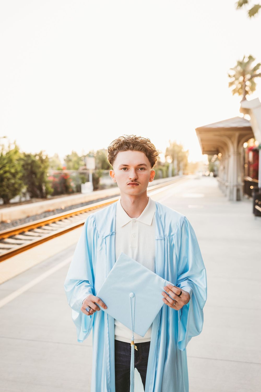 Young graduate in light blue cap and gown holding mortarboard at train station platform during golden hour.