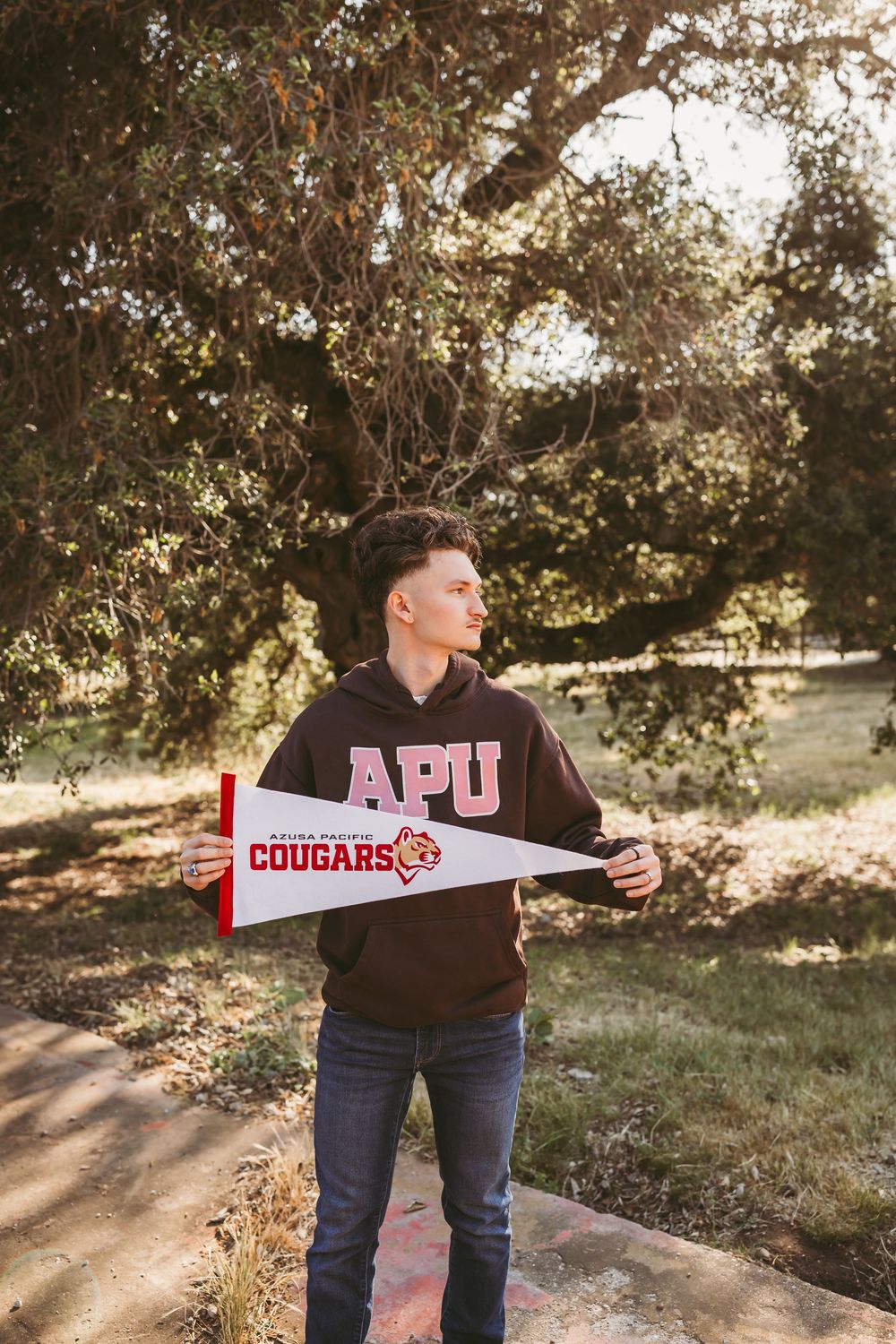 Young man holding an APU Cougars pennant flag outdoors under large oak trees.