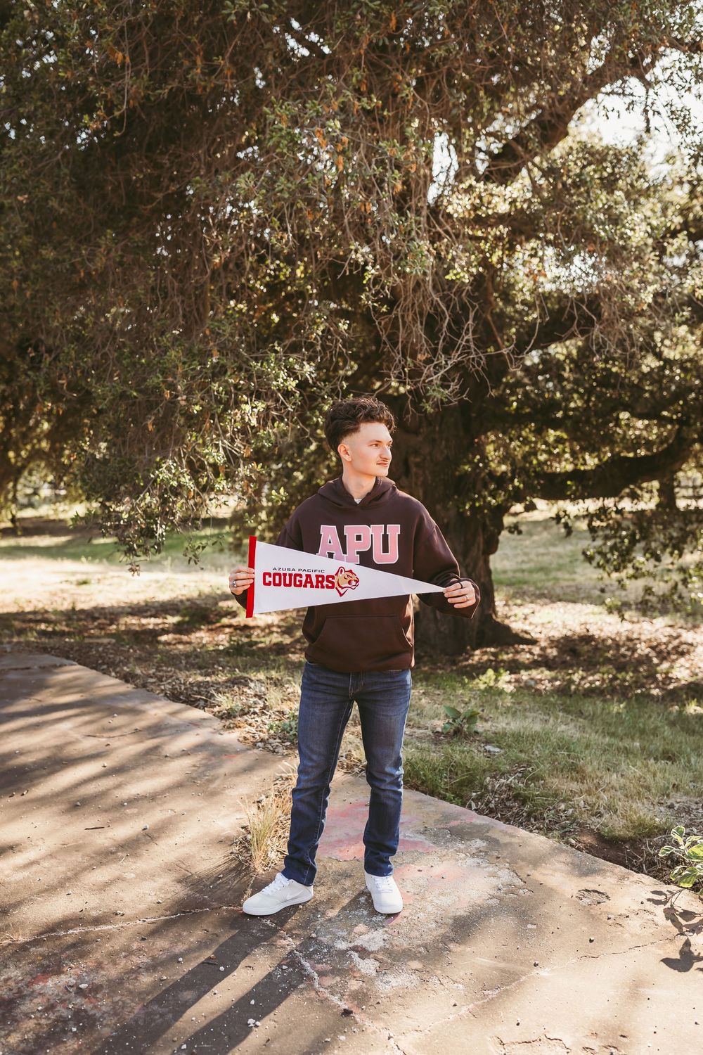 Young man in APU Cougars hoodie holding a pennant flag on a sunlit outdoor path.