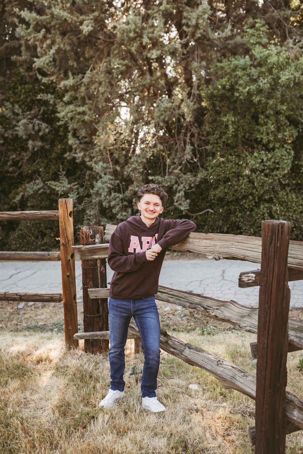 Smiling teen in APU hoodie leaning on rustic wooden fence surrounded by trees.