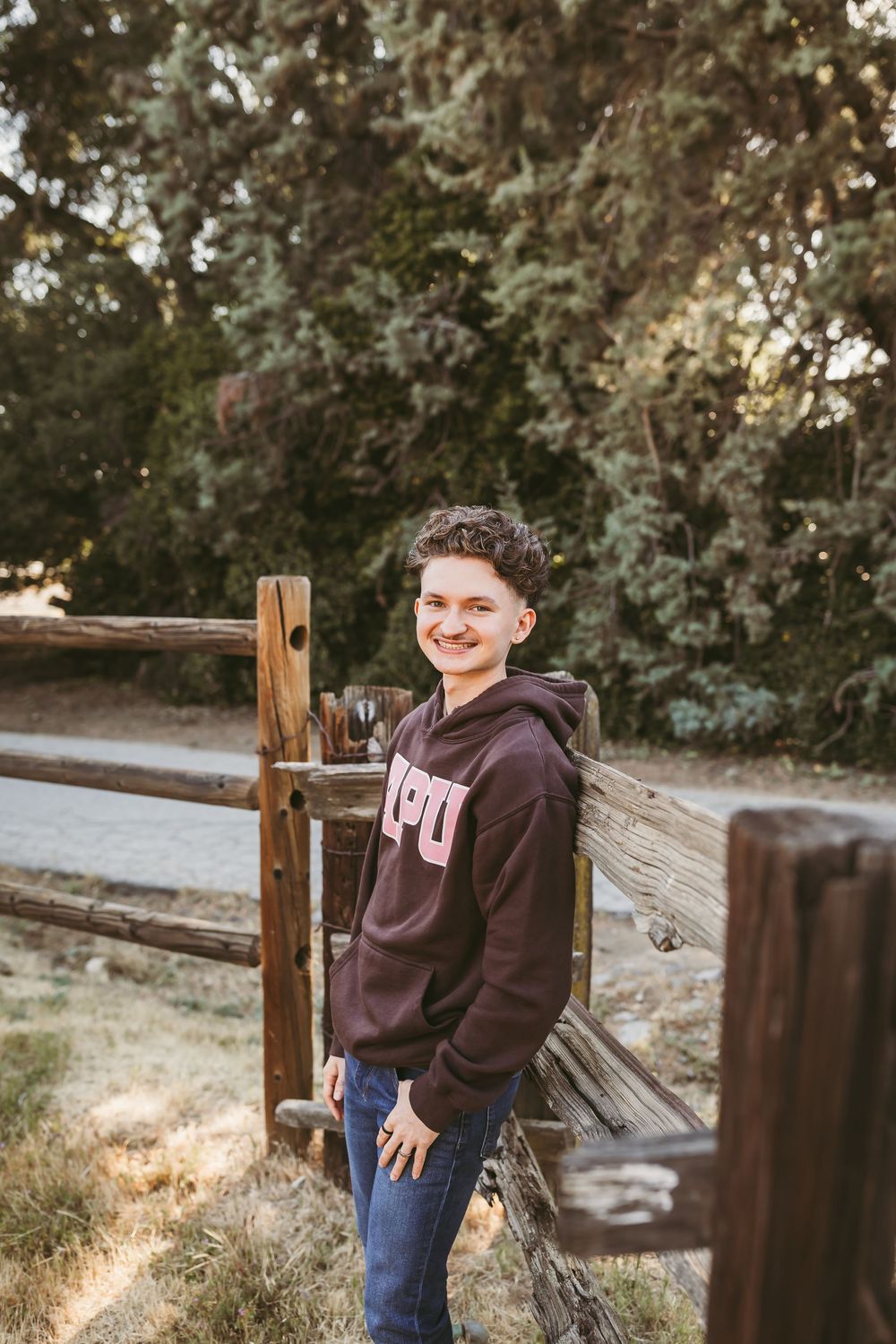 Smiling teen in maroon hoodie leaning against rustic wooden fence outdoors surrounded by trees.