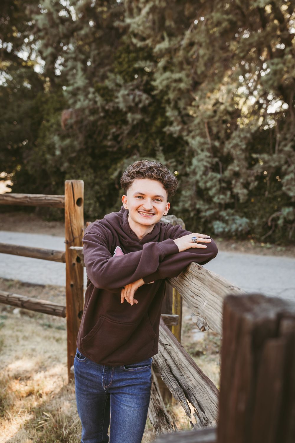 Smiling young man in maroon hoodie leaning on rustic wooden fence surrounded by lush green trees.