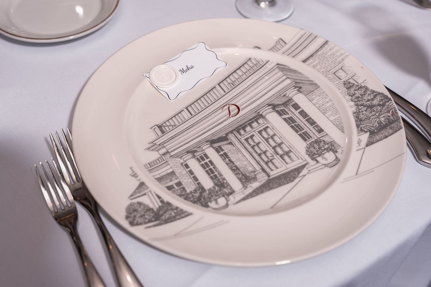 Elegant place setting with decorative plate featuring, silverware, and name card on white tablecloth. Deerfield Golf Club Newark De