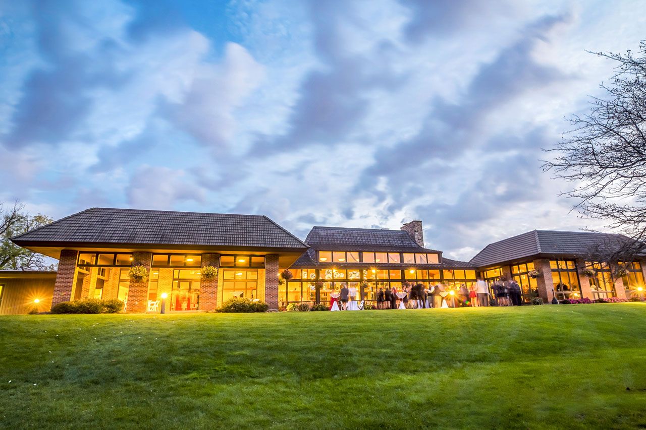 Outdoor wedding ceremony setup with white folding chairs on green lawn, floral arch, and serene pond backdrop.