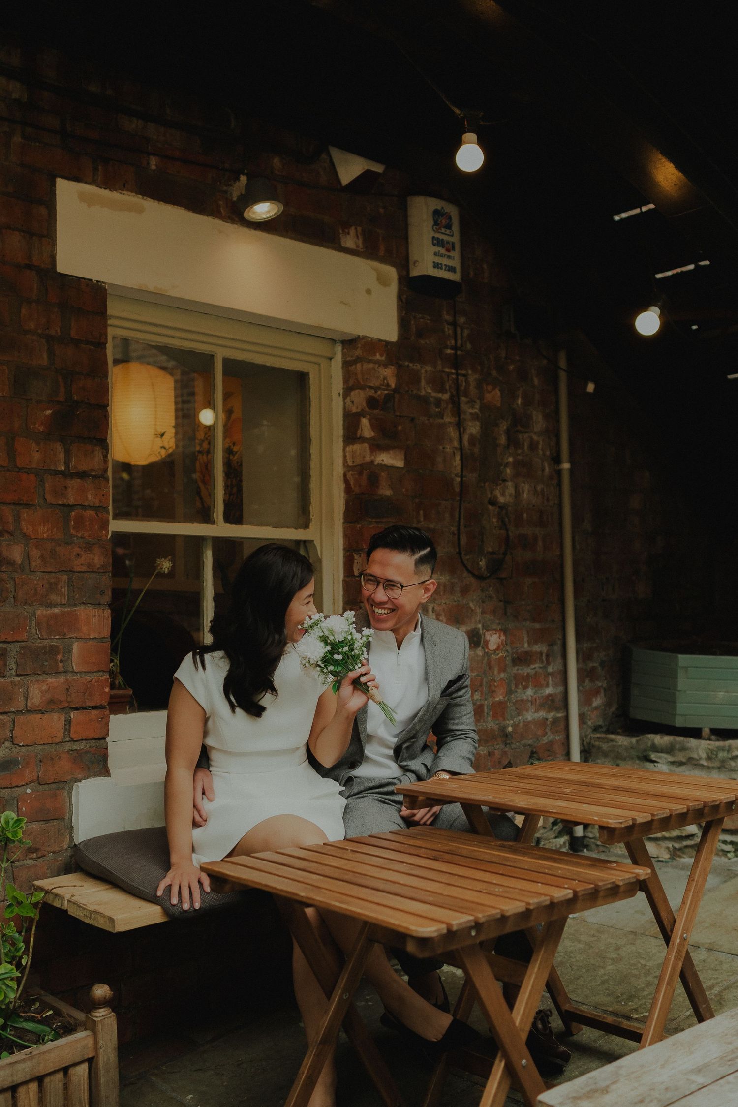 Couple sitting together at a wooden cafe table against a brick wall, woman holding a small bouquet, sharing a romantic moment.