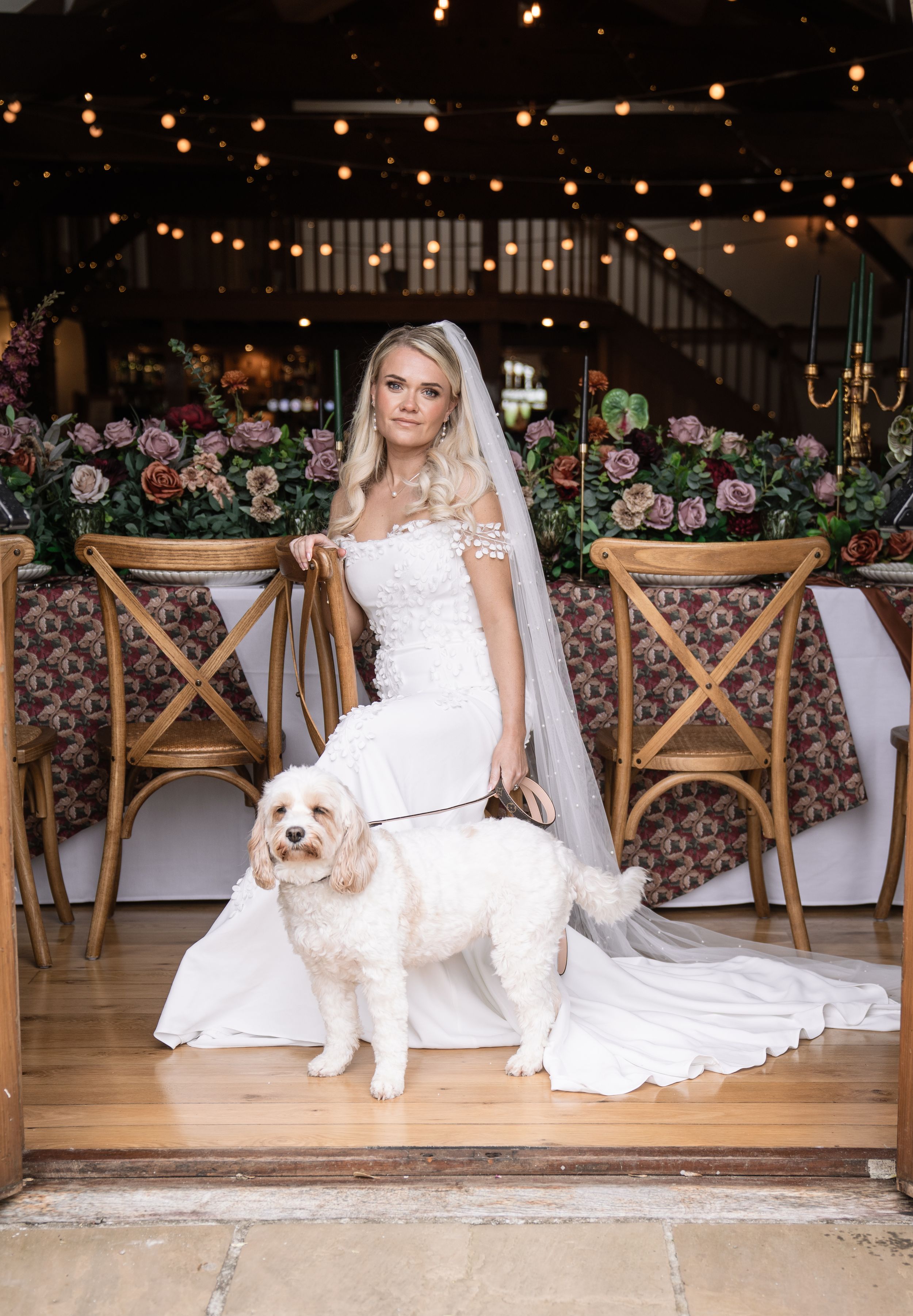 Bride in white lace gown and veil poses with fluffy white dog in elegant floral wedding venue with string lights.