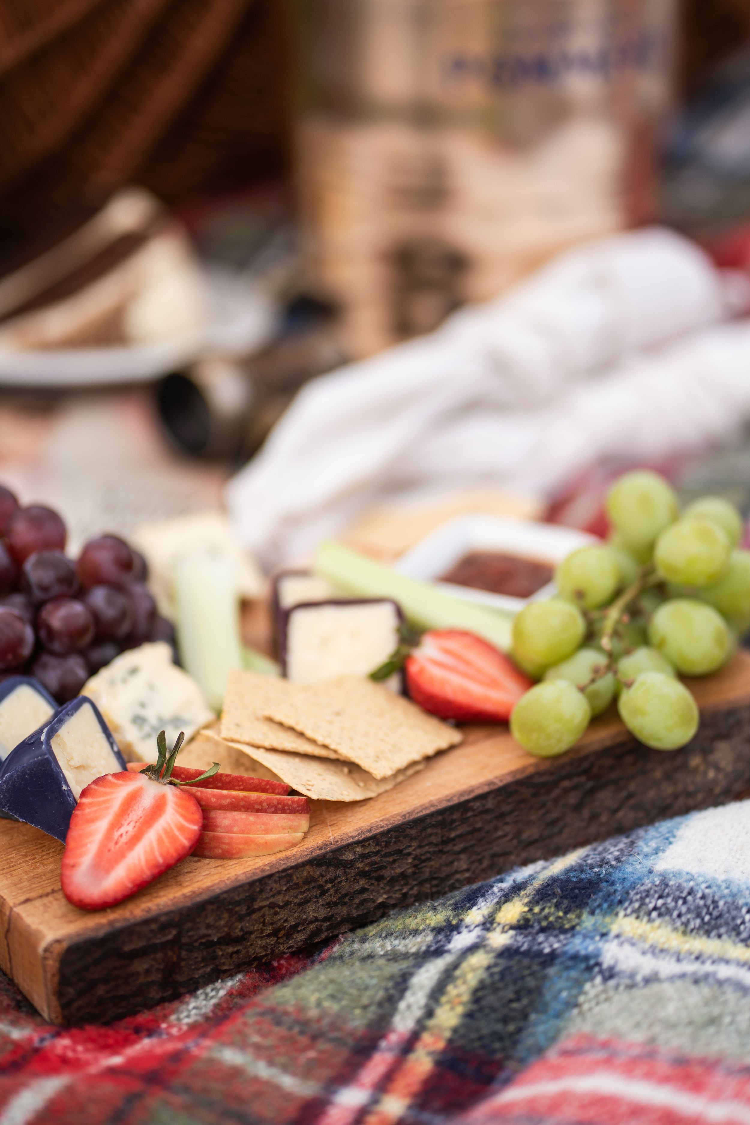 Rustic wooden charcuterie board with cheese, crackers, strawberries, and grapes on a plaid picnic blanket.