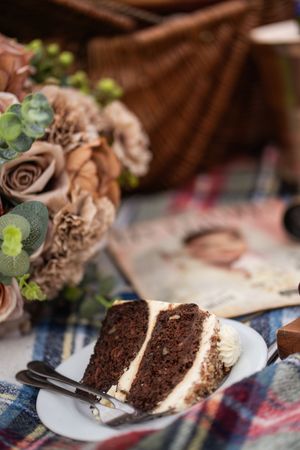 A slice of layered chocolate cake with cream frosting on a white plate beside a bouquet of roses at a picnic setting.
