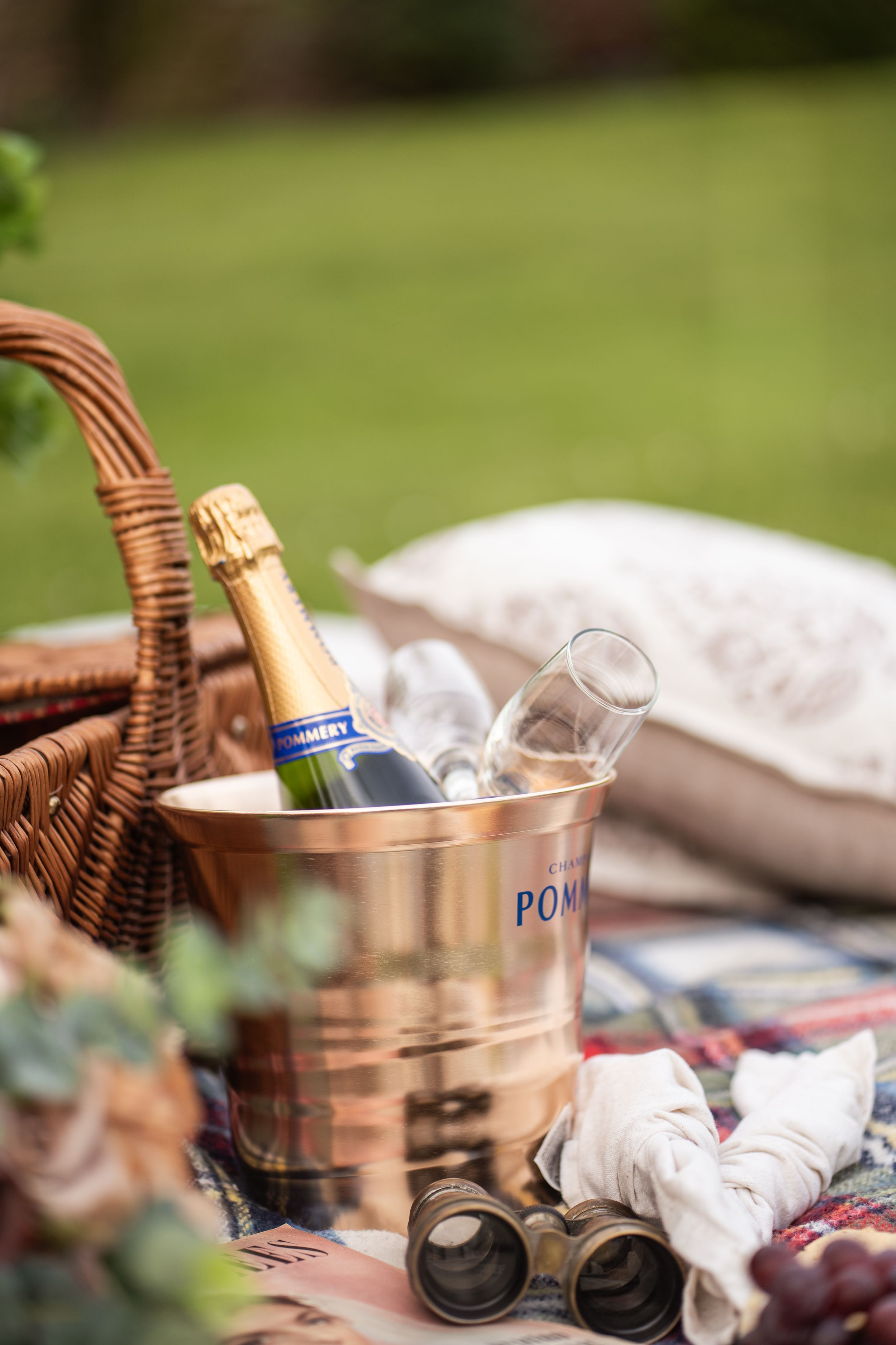 Champagne bottle chilling in copper ice bucket with glasses beside wicker picnic basket on blanket outdoors.
