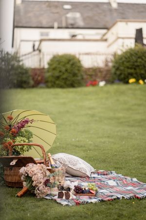A charming garden picnic setup with a plaid blanket, wicker basket, parasol, flowers, and champagne on lush green grass.