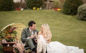 Bride and groom sitting on grass sharing a romantic picnic with flowers and umbrella on their wedding day.