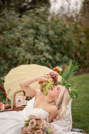 Bride in lace gown lying on a picnic blanket eating grapes, surrounded by flowers and a yellow umbrella in a garden.