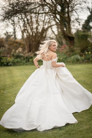 A blonde bride twirls in a stunning off-shoulder white ballgown wedding dress in a lush garden setting.