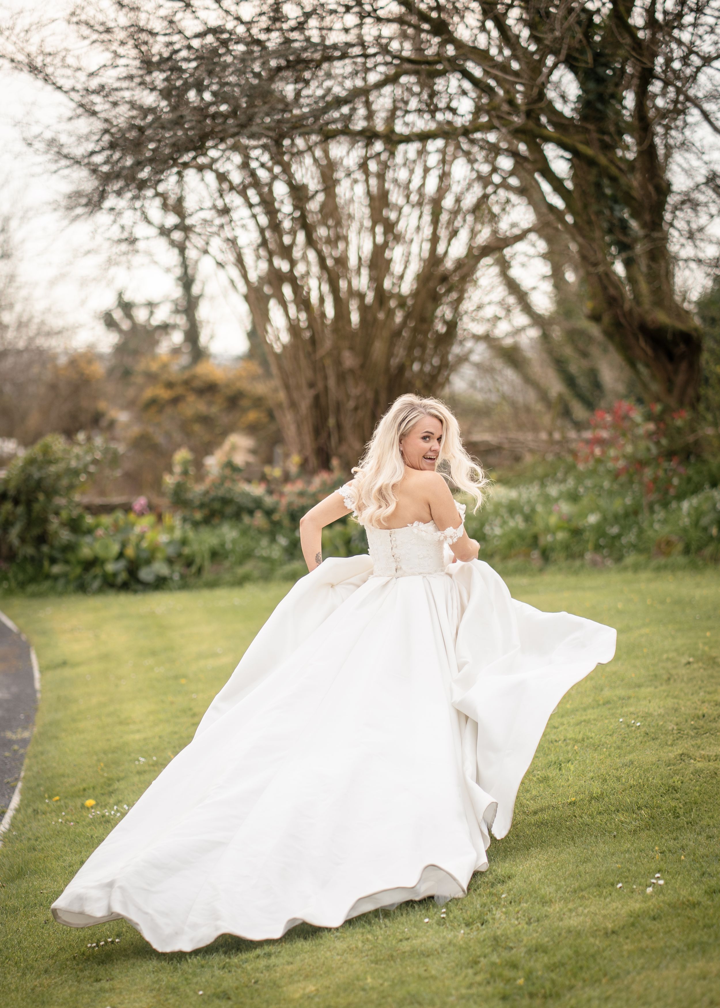 Blonde bride in a flowing off-shoulder white ball gown twirling in a lush garden with bare trees.