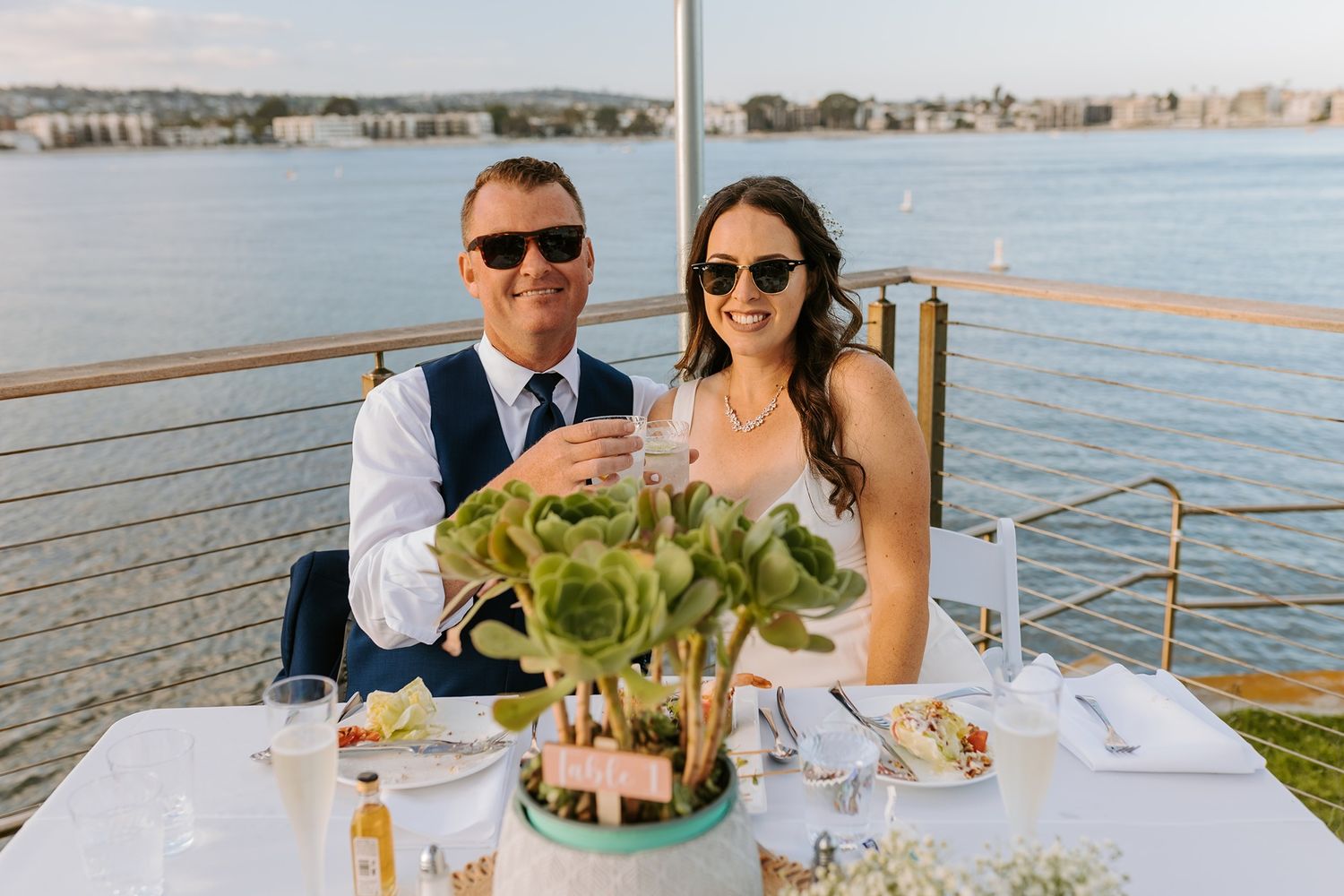 Couple in sunglasses toasting champagne at outdoor waterfront wedding reception table with greenery centerpiece at Tower Beach Club.