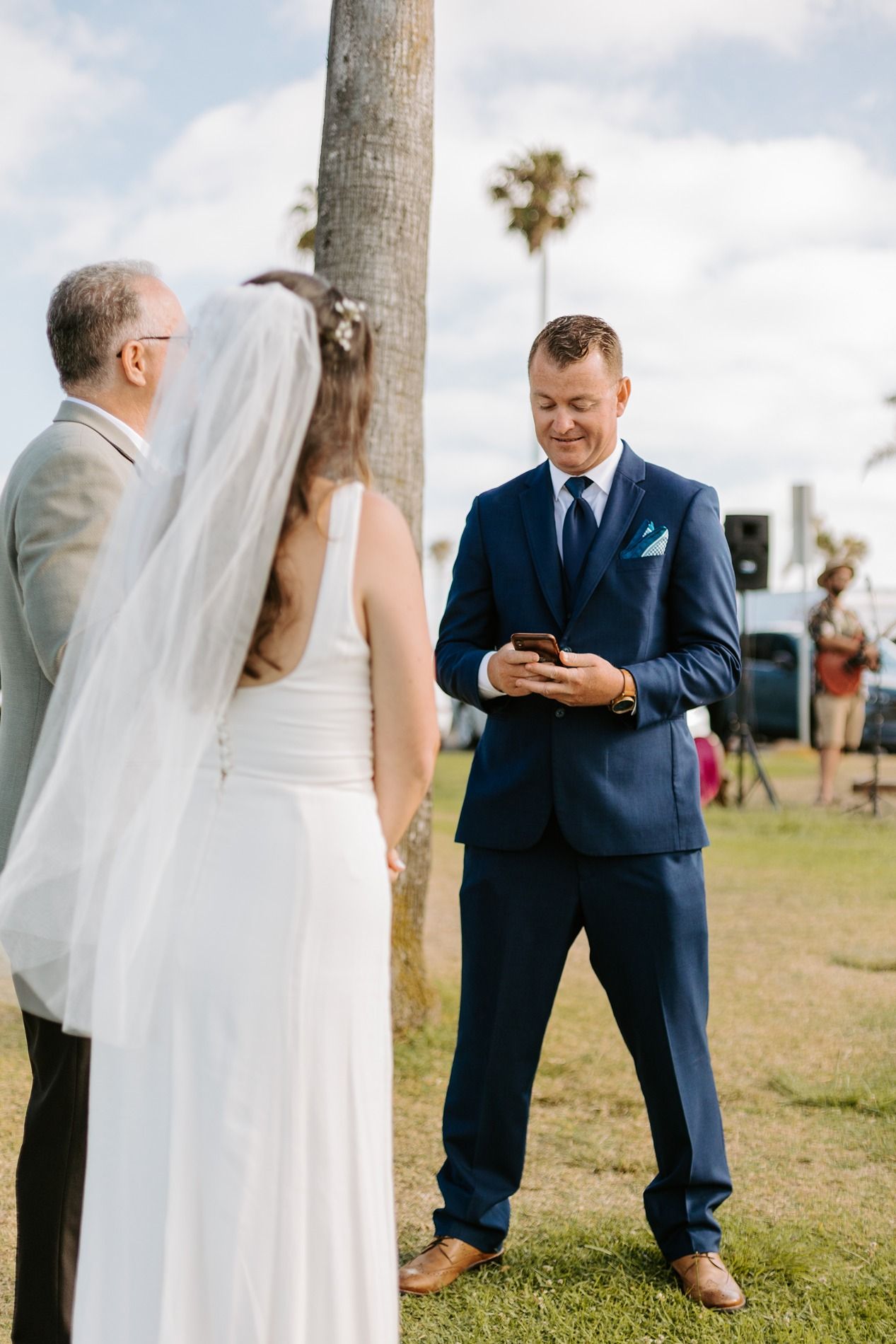 Groom in navy suit reads vows from phone while bride in white dress and veil stands with officiant outdoors at Tower Beach Club.