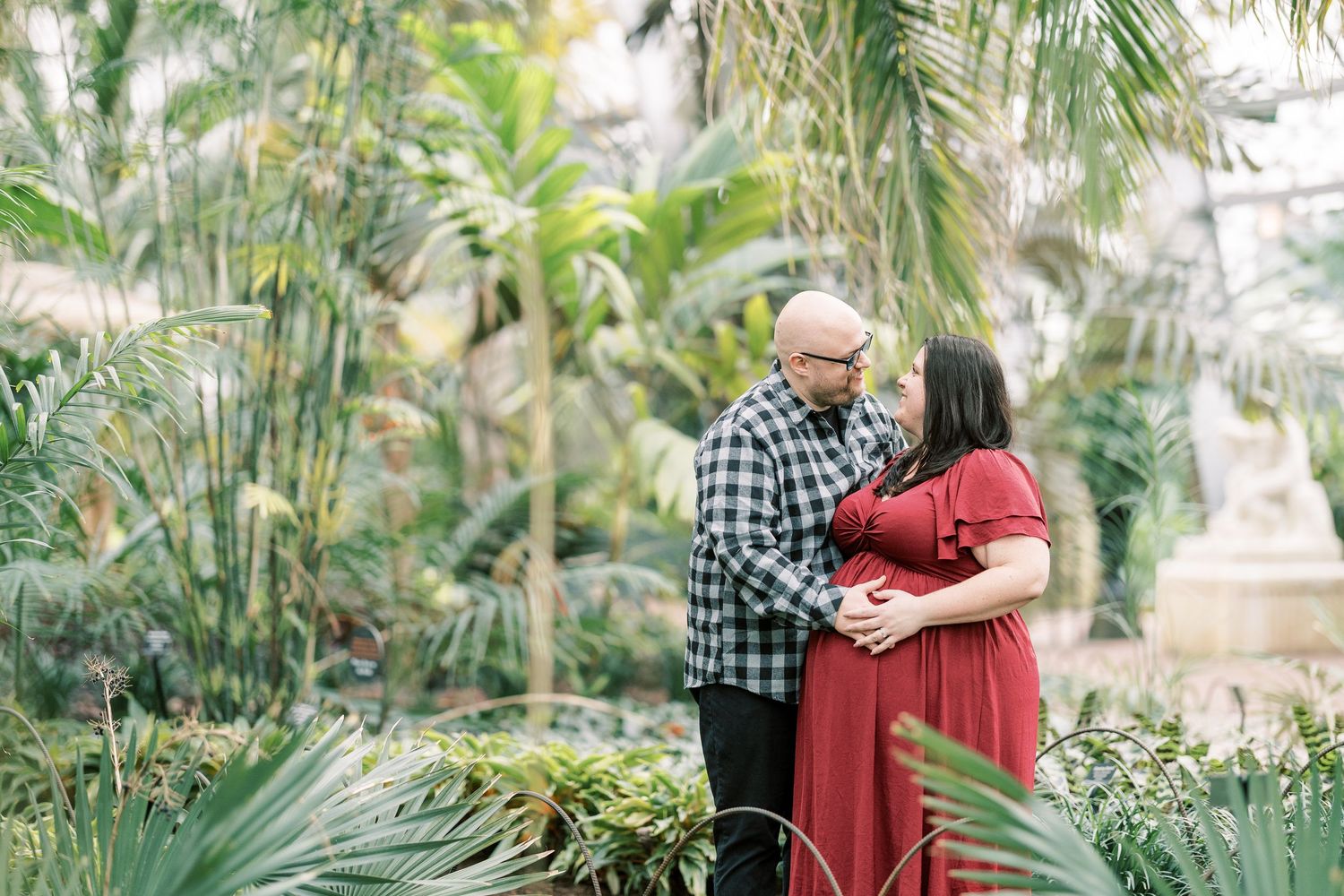 Couple sharing romantic moment in lush tropical greenhouse, woman in red maternity dress.