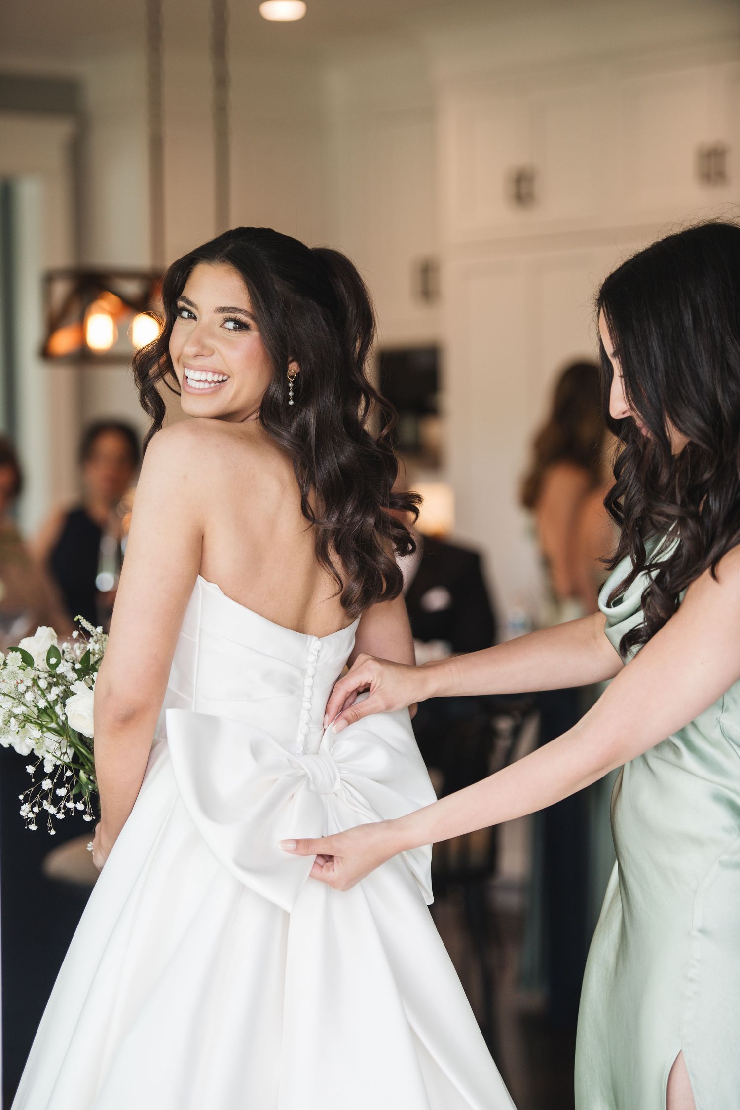 Smiling bride in strapless white gown with bow back being buttoned by bridesmaid in sage green dress.