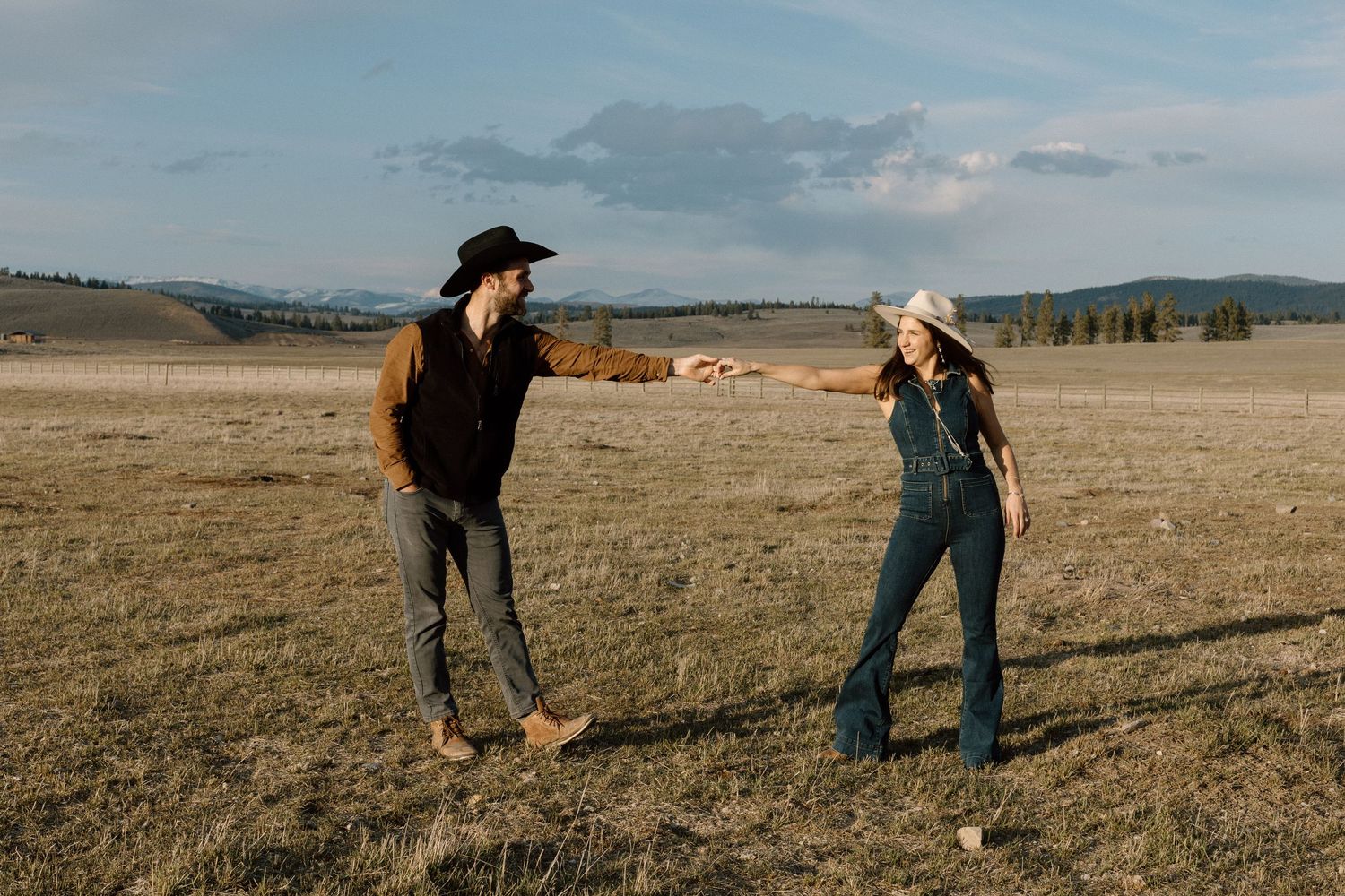 A couple in western attire reaching toward each other in an open field with mountains in the background.