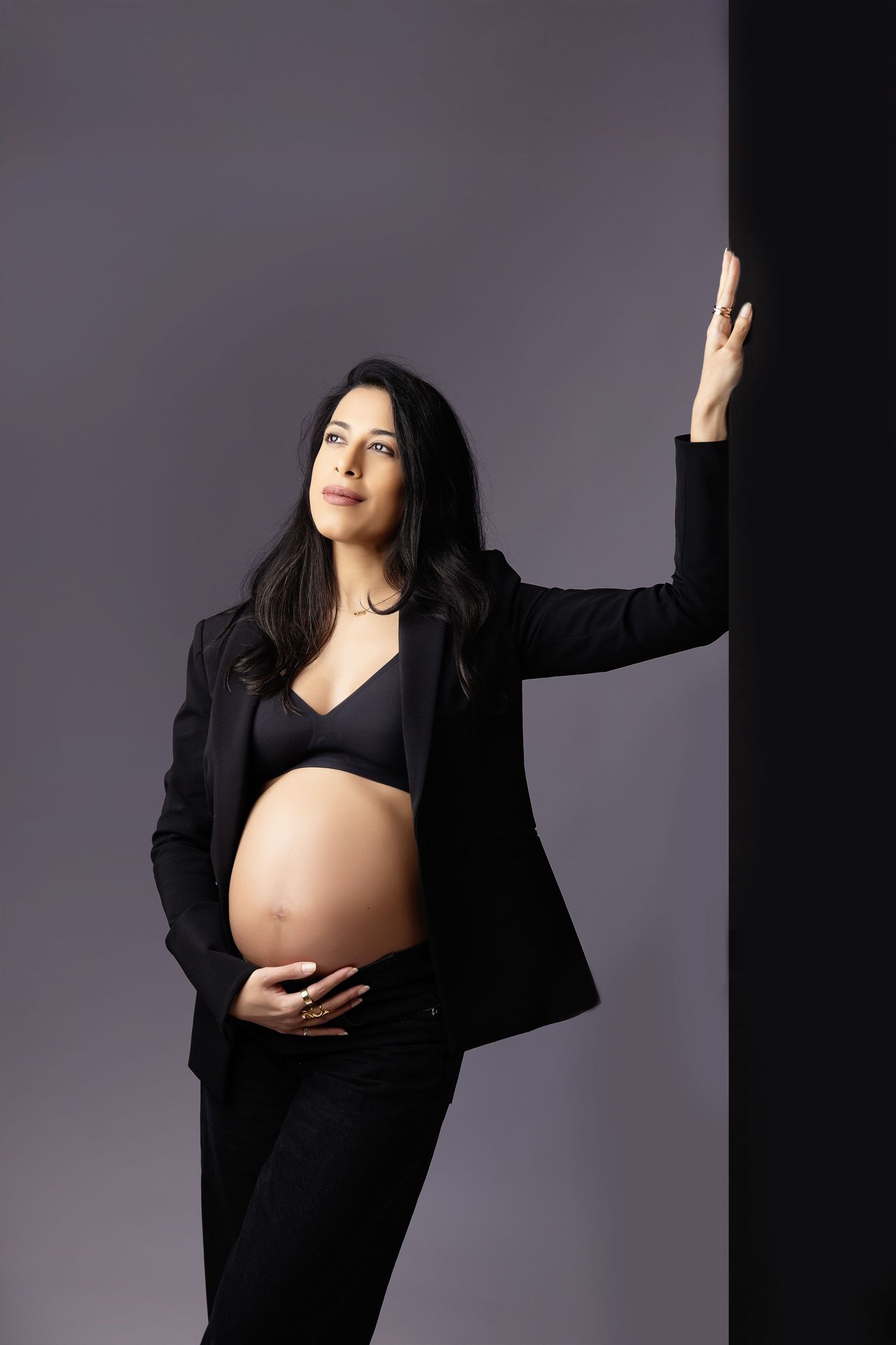 Pregnant woman in black blazer and bra cradling baby bump, leaning against wall in studio portrait.