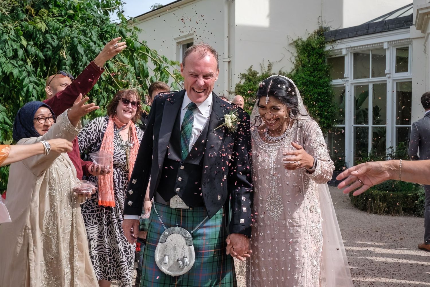 Bride and groom walk through confetti shower at outdoor wedding, groom in tartan kilt, bride in embroidered gown.