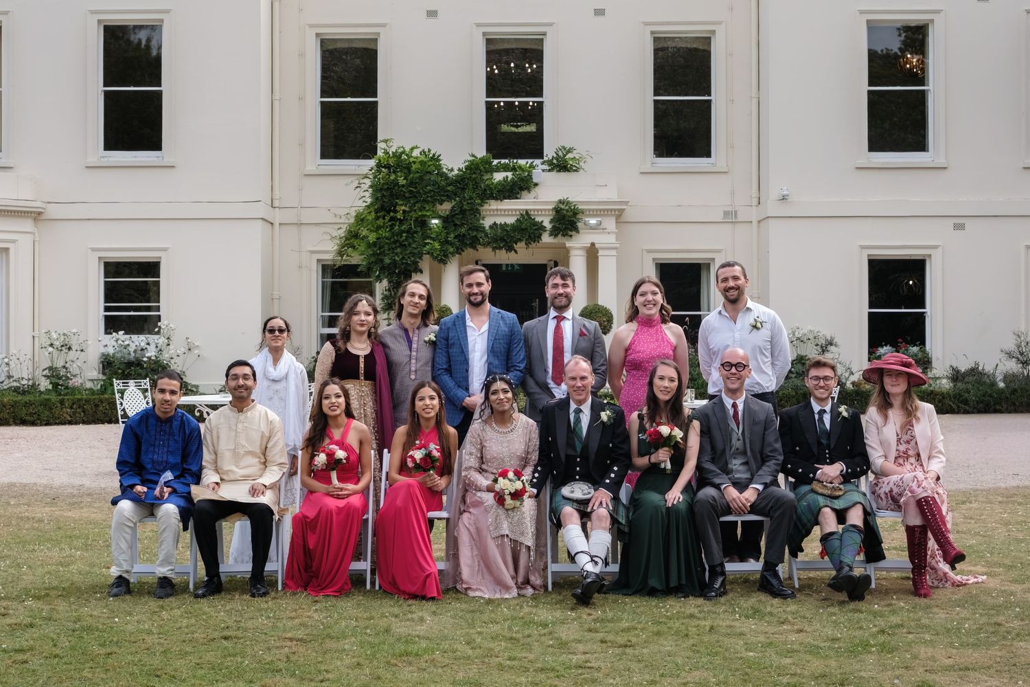 Wedding group photo with bride, groom, and guests posing in front of an elegant white manor house.