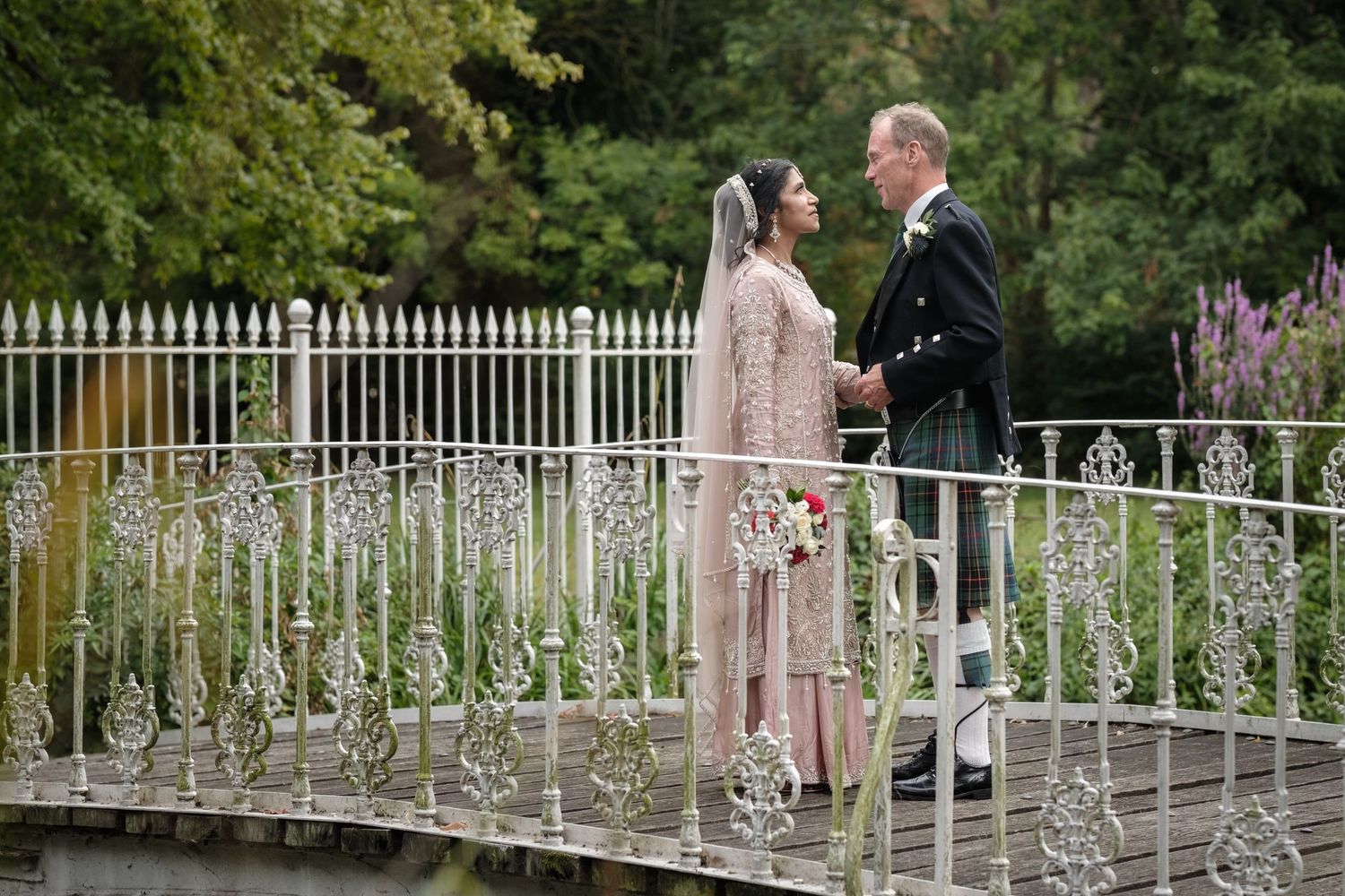 Bride and groom in Scottish kilt share a romantic moment on an ornate white bridge in a lush garden setting.