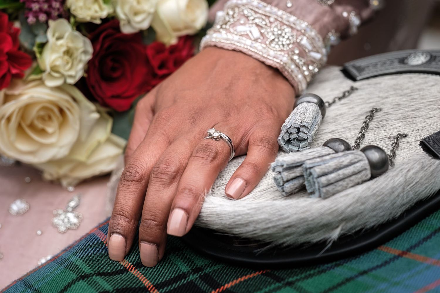 Close-up of a bride's hand with engagement ring, floral bouquet, tartan fabric, and Scottish sporran at a wedding.