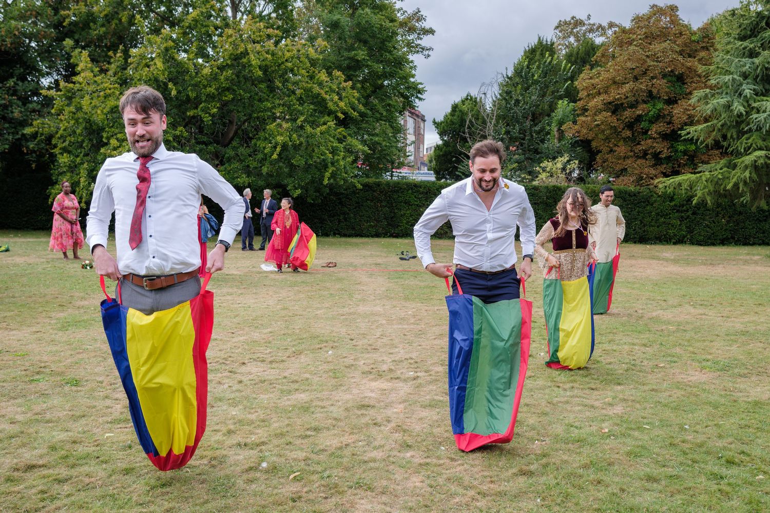 Two men compete in a colorful sack race on a green lawn at an outdoor garden party event.
