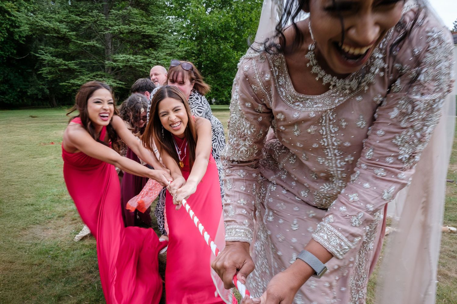 Bridesmaids in red saris playfully tug a rope with a bride in embroidered gown during outdoor wedding celebration.