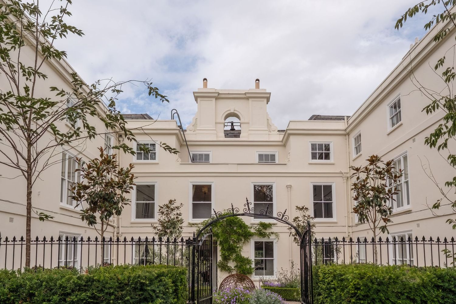 Elegant white Georgian townhouse with ornate iron gates, manicured gardens, and classic architectural details under a cloudy sky.