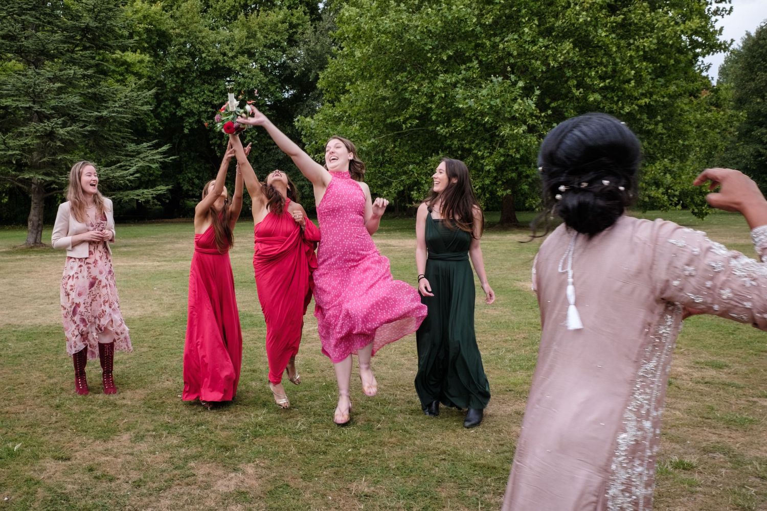 Bridesmaids in colorful dresses reaching for bouquet toss in garden.