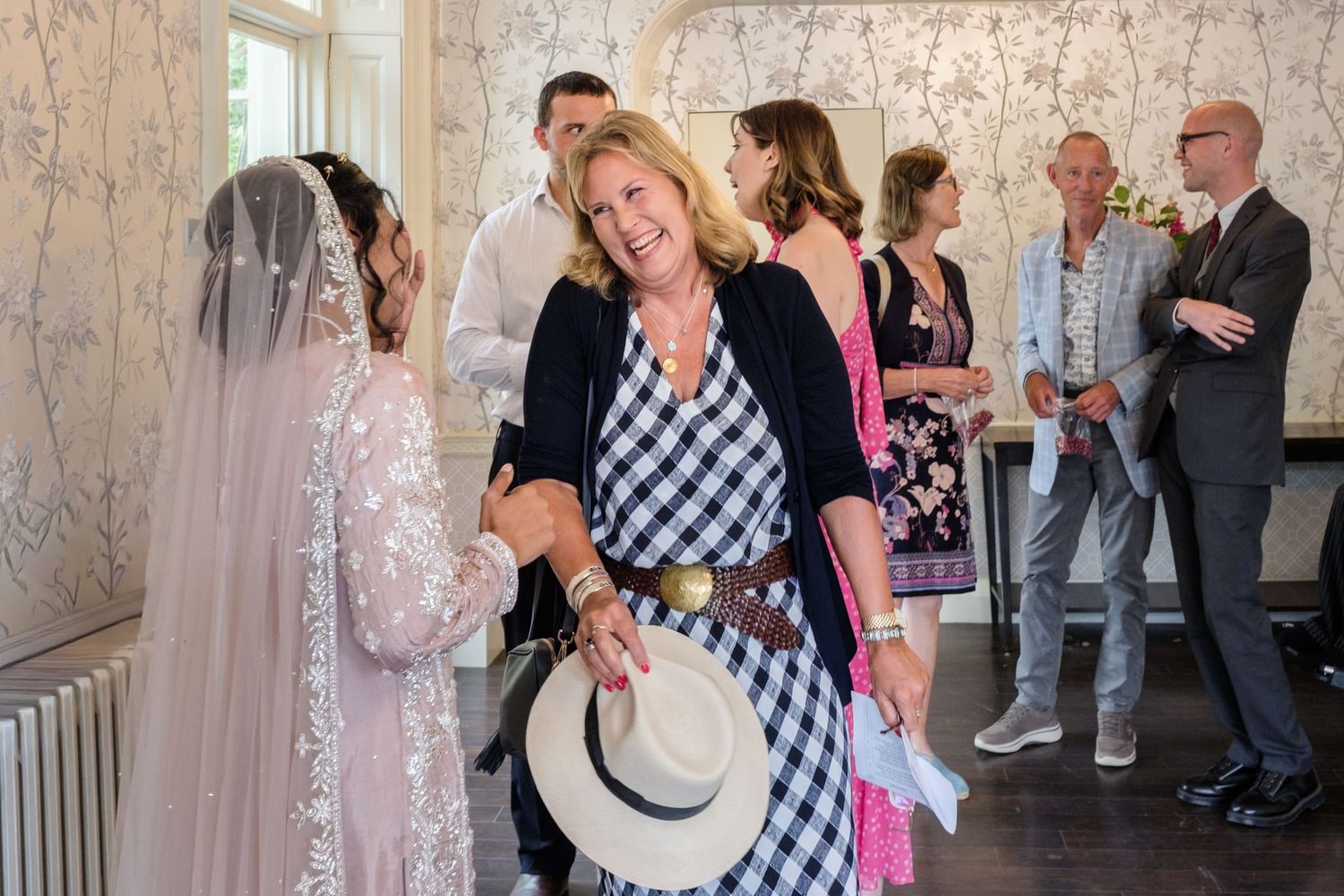 Woman in checkered dress holding white hat smiles at bride in veil at elegant indoor wedding reception.