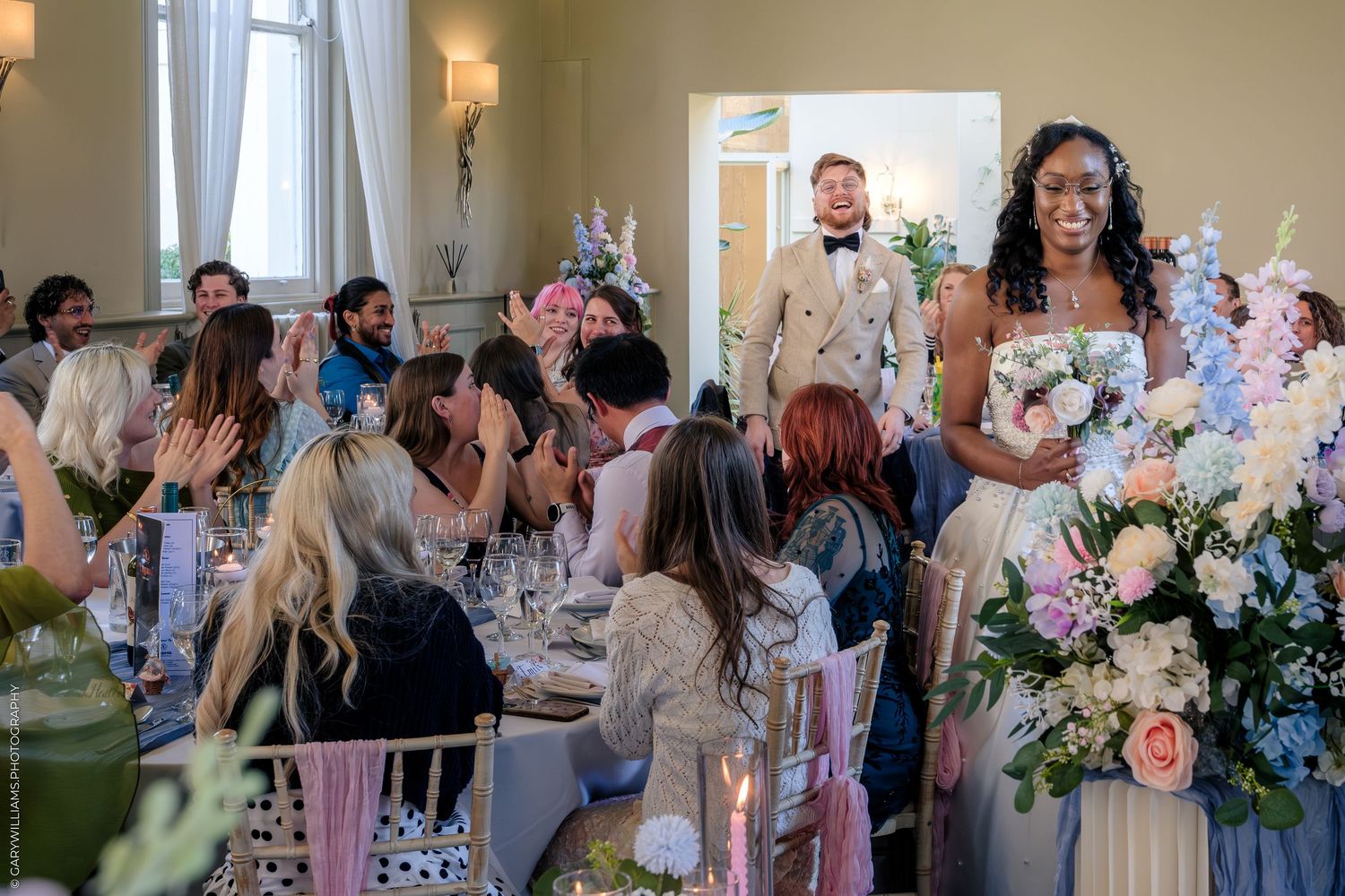 Bride holding bouquet stands before seated wedding guests at elegant floral-decorated reception table in bright venue.