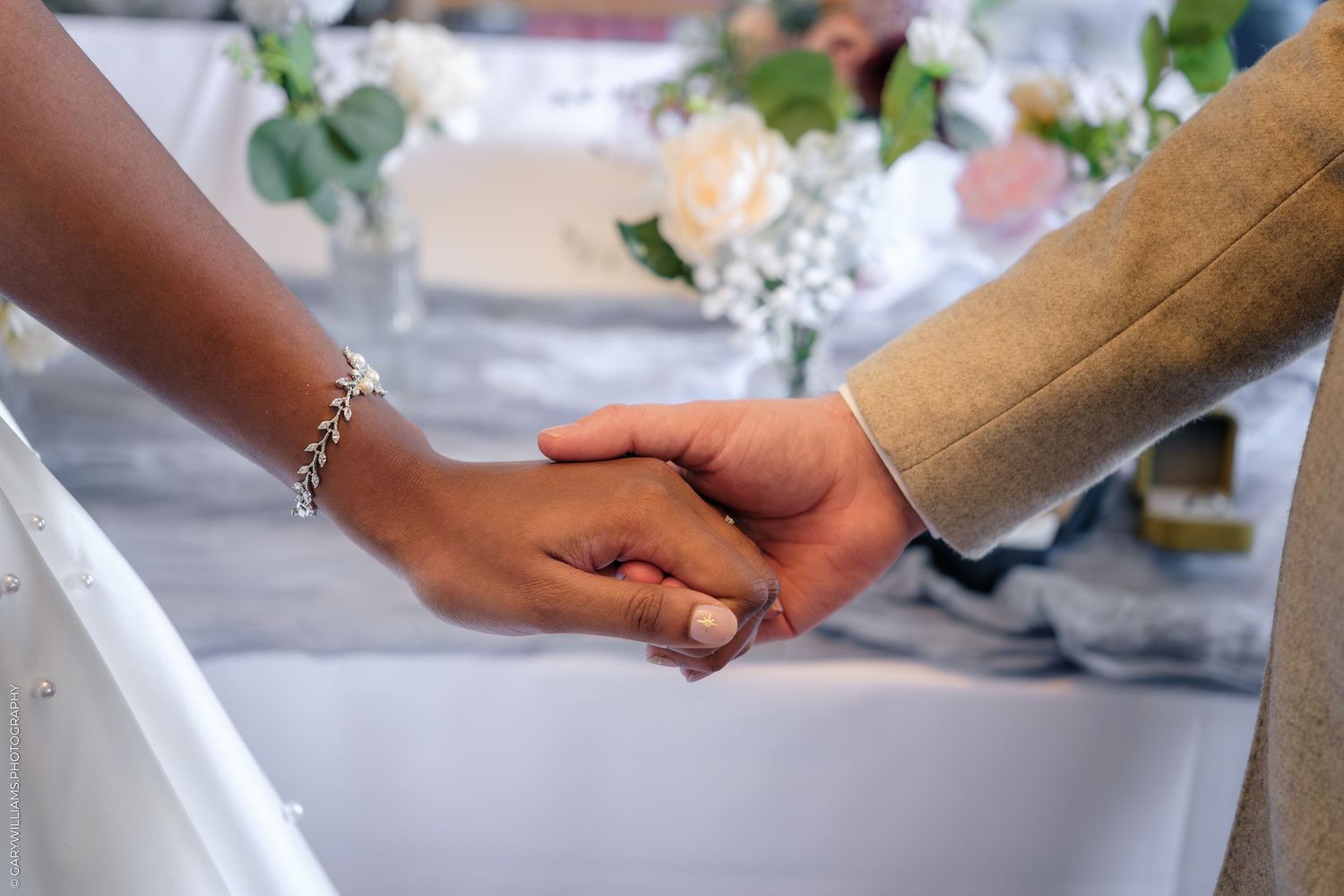 Interracial couple holding hands at a wedding ceremony, bride wearing a silver bracelet and engagement ring.