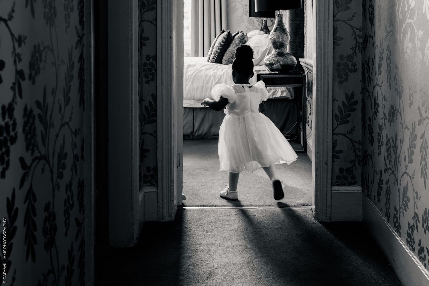 Black and white photo of a young girl in a white dress running down a hallway toward a bedroom.