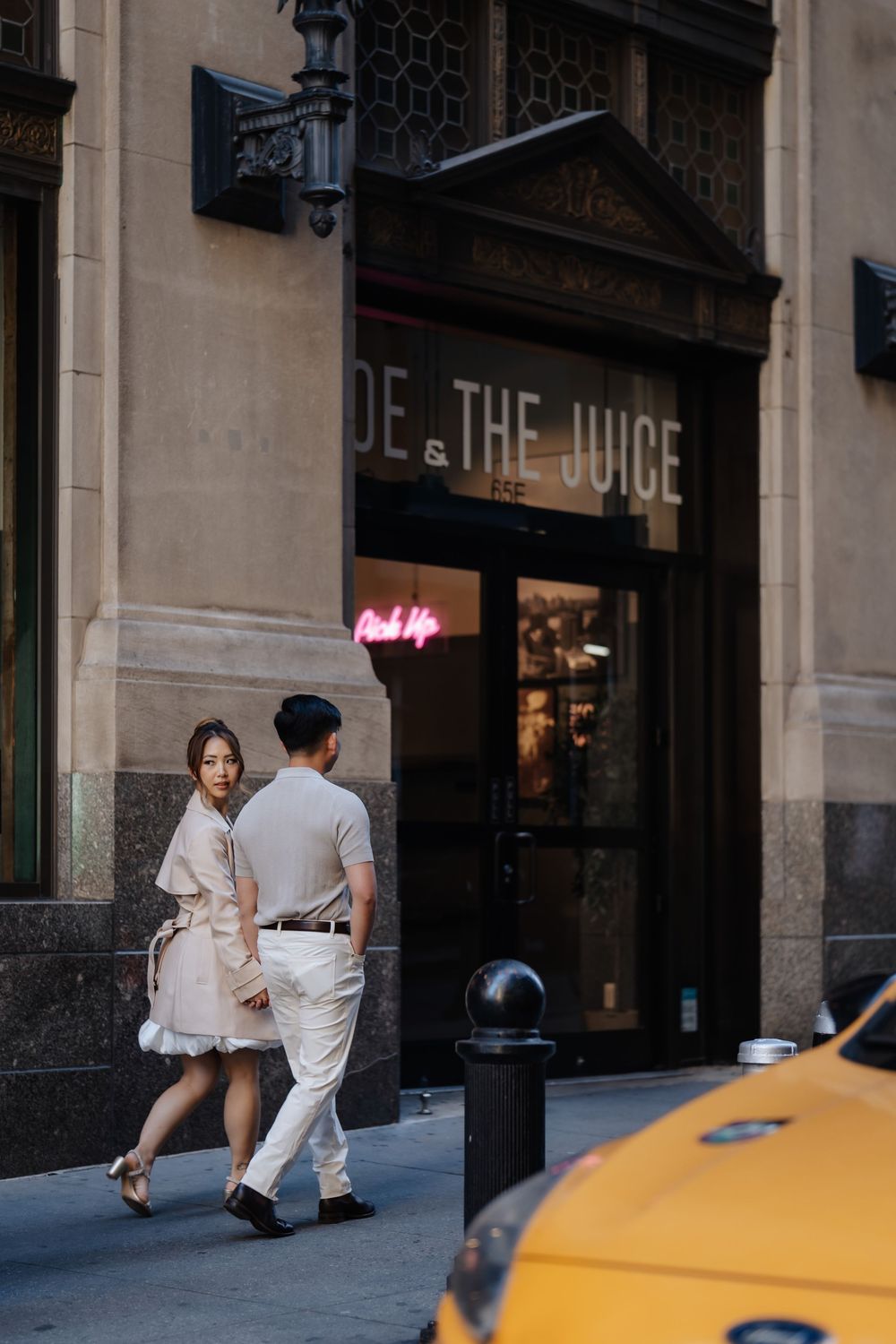 Couple walking past Joe & The Juice cafe on a sunny urban street corner with a yellow taxi nearby.
