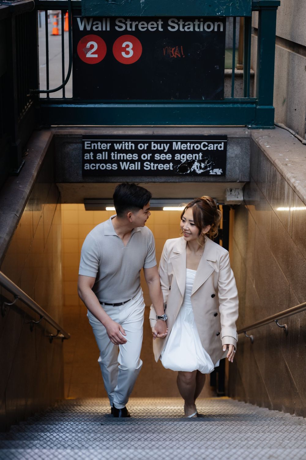 Couple holding hands walking up Wall Street Station subway steps in NYC, man in gray polo and woman in beige blazer.