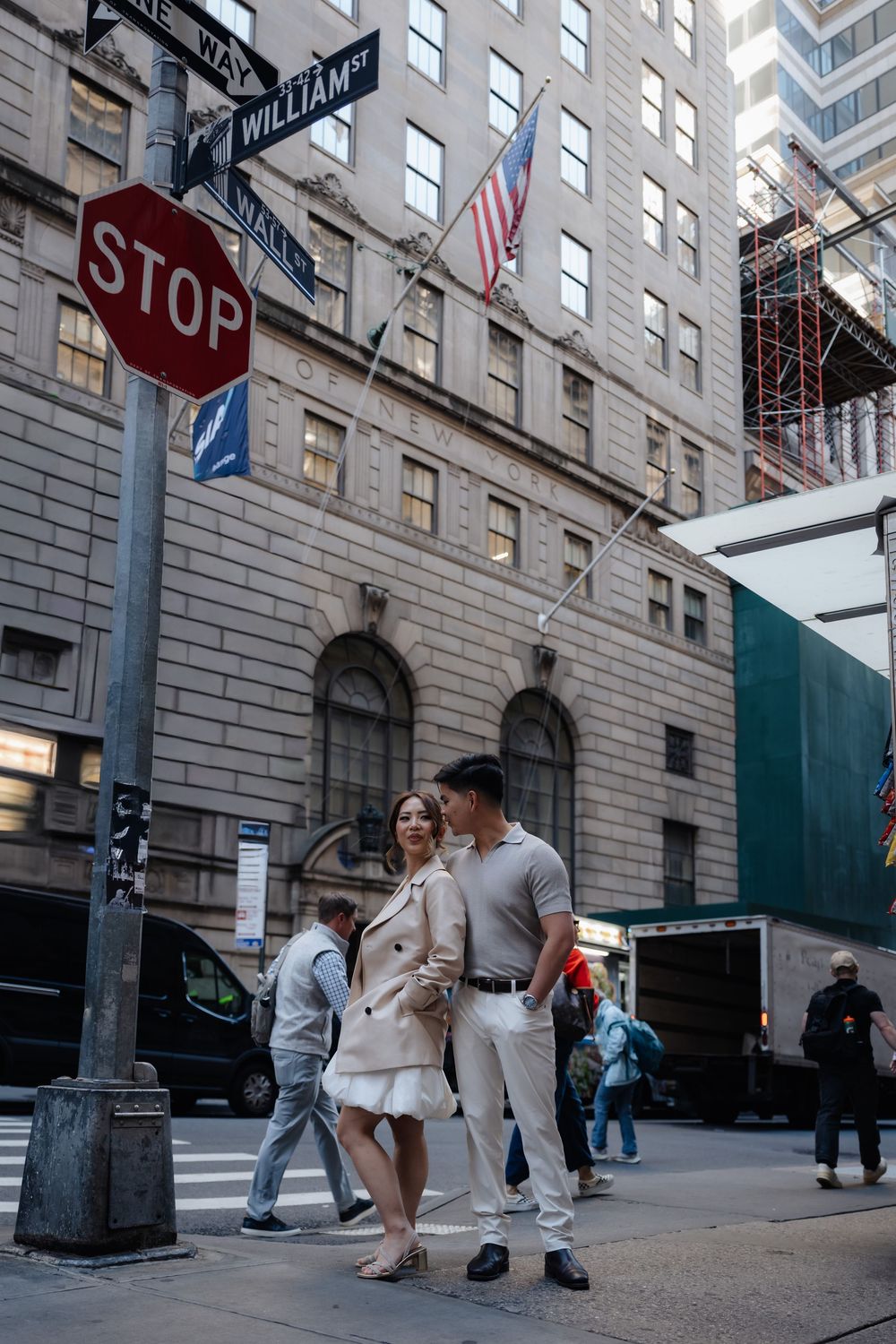 Stylish couple posing at William Street intersection in New York City with a stop sign and American flag backdrop.