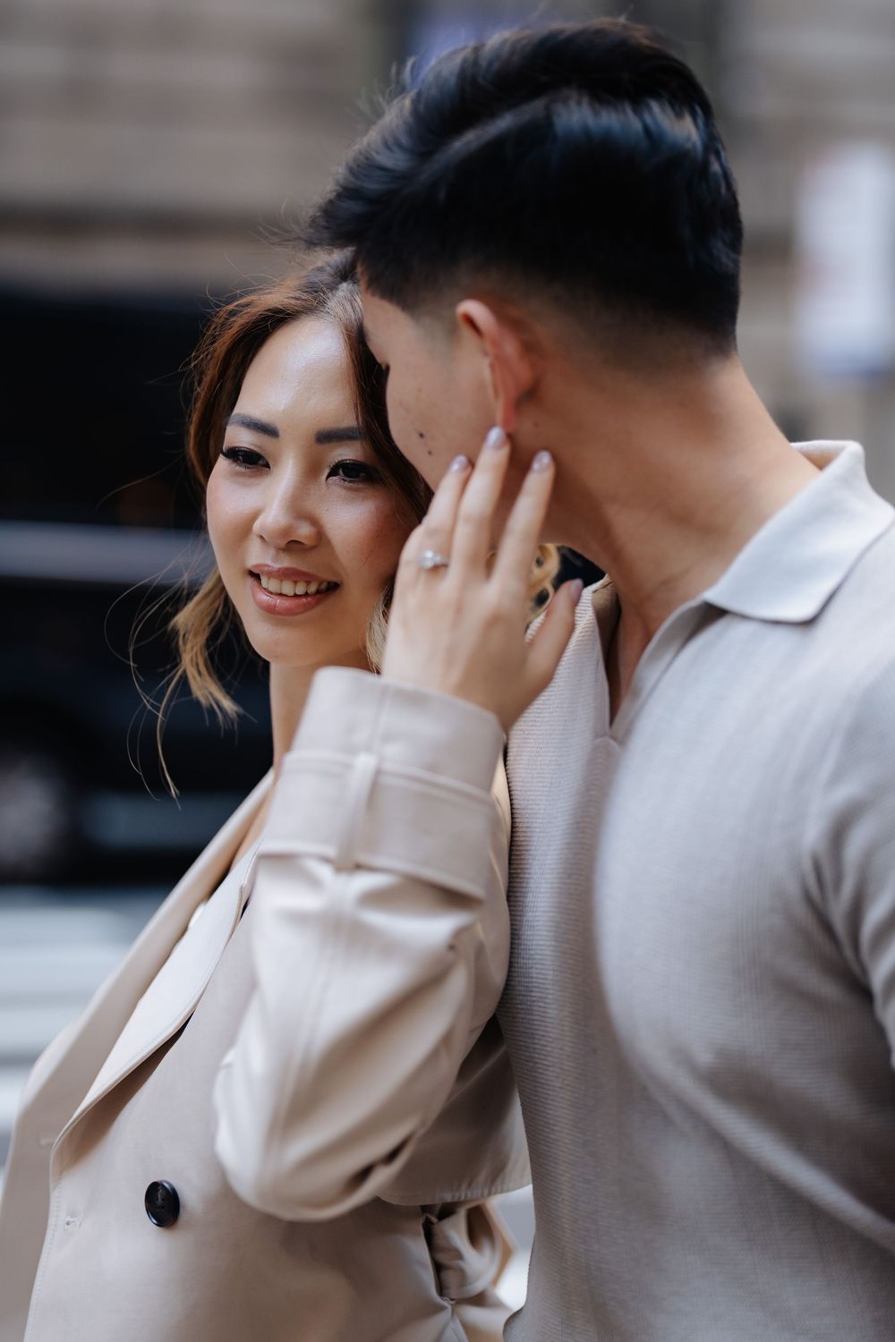 A smiling Asian woman in a beige blazer tenderly touches a man's face, showcasing an engagement ring outdoors.