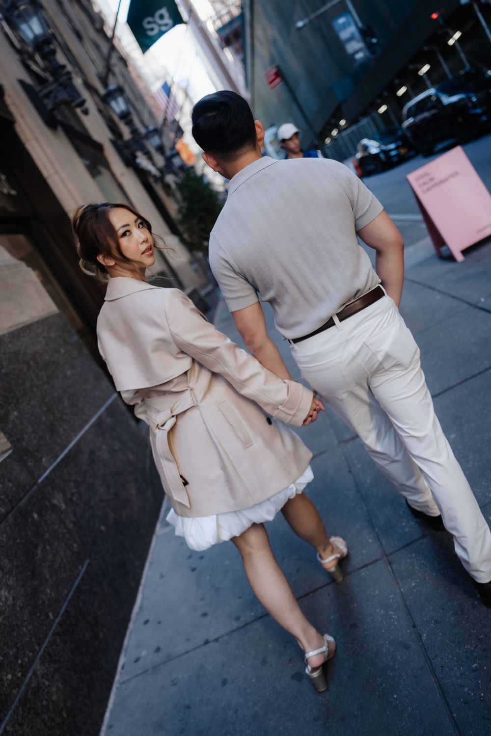 Couple holding hands walking on a city sidewalk, woman in beige trench coat glancing back over her shoulder.