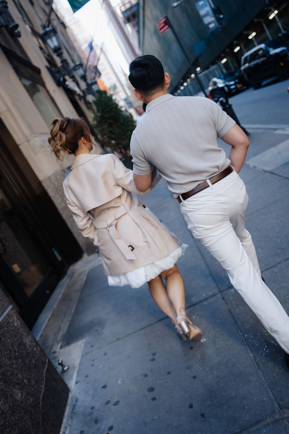 Couple walking hand in hand on a city sidewalk, wearing coordinated beige and white outfits in an urban setting.