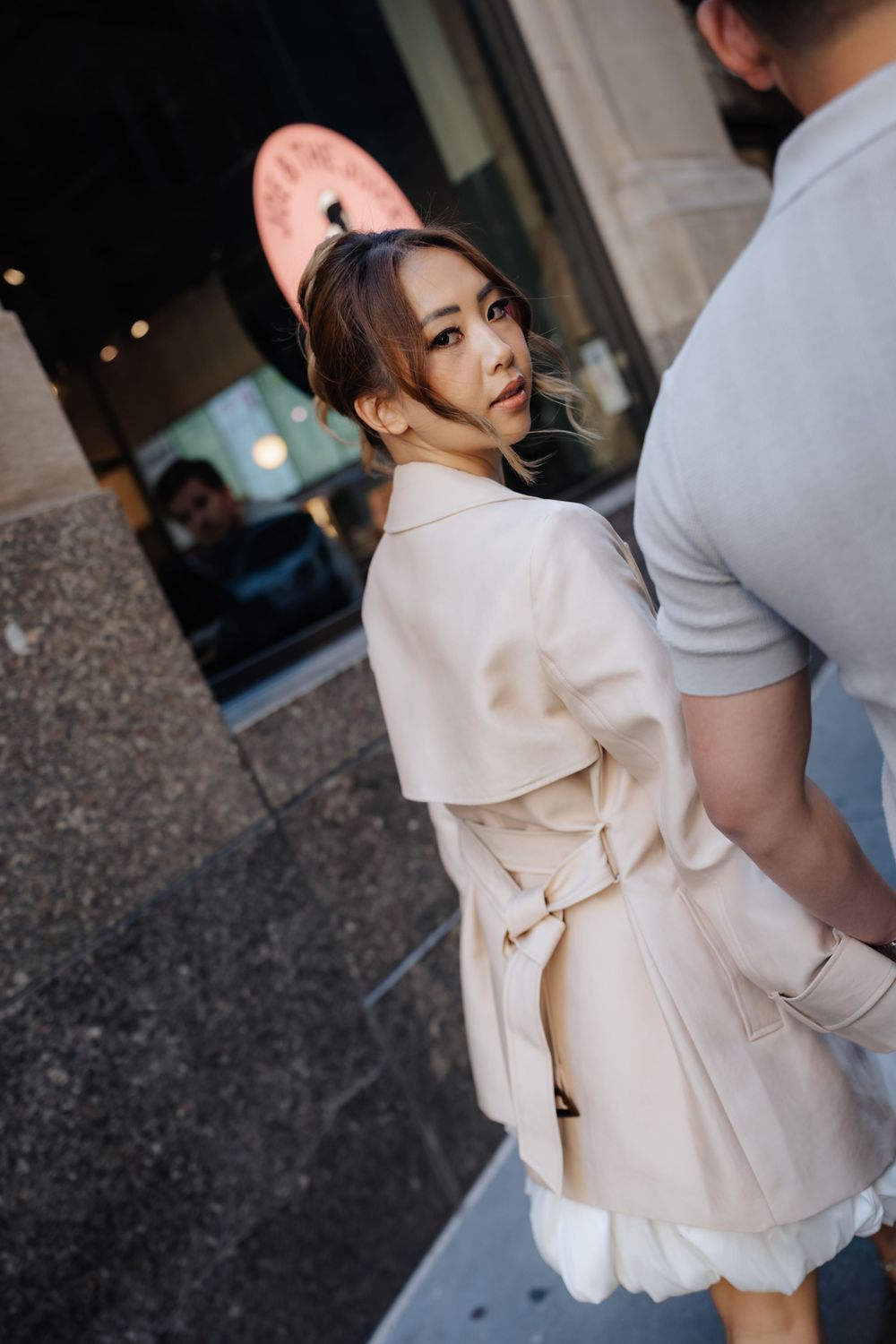 Woman in beige trench coat glances back over shoulder on urban sidewalk.