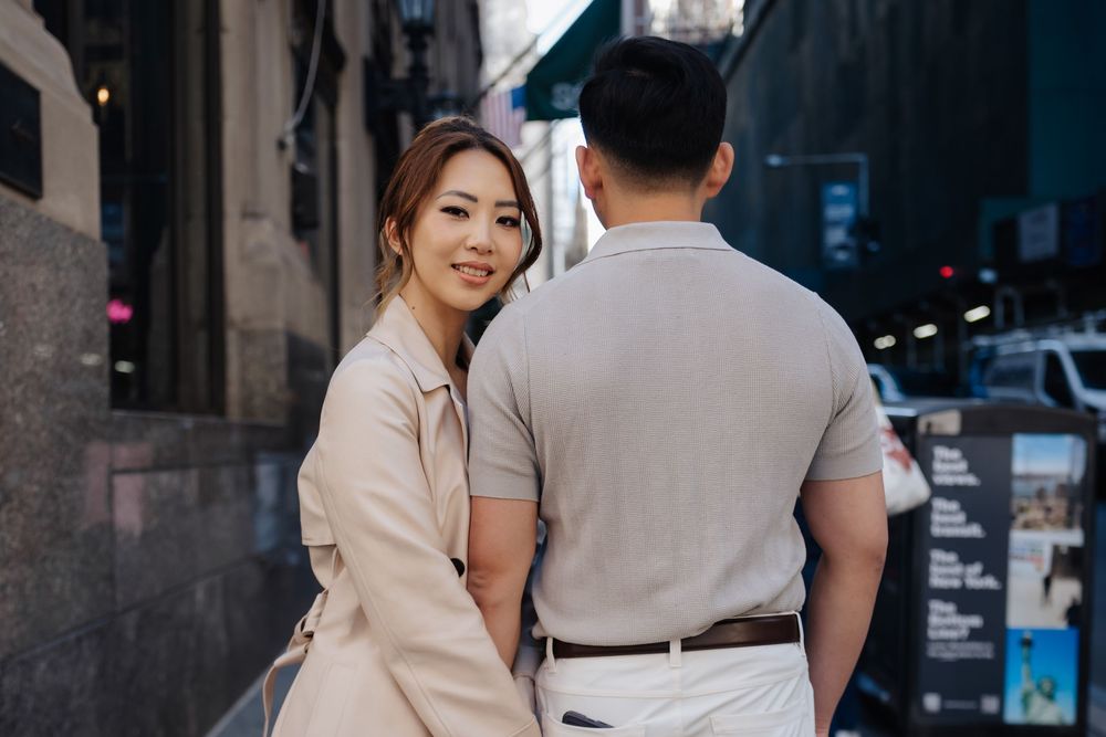 Asian couple posing on a city street, woman in pink coat smiling over shoulder, man in beige polo shirt.