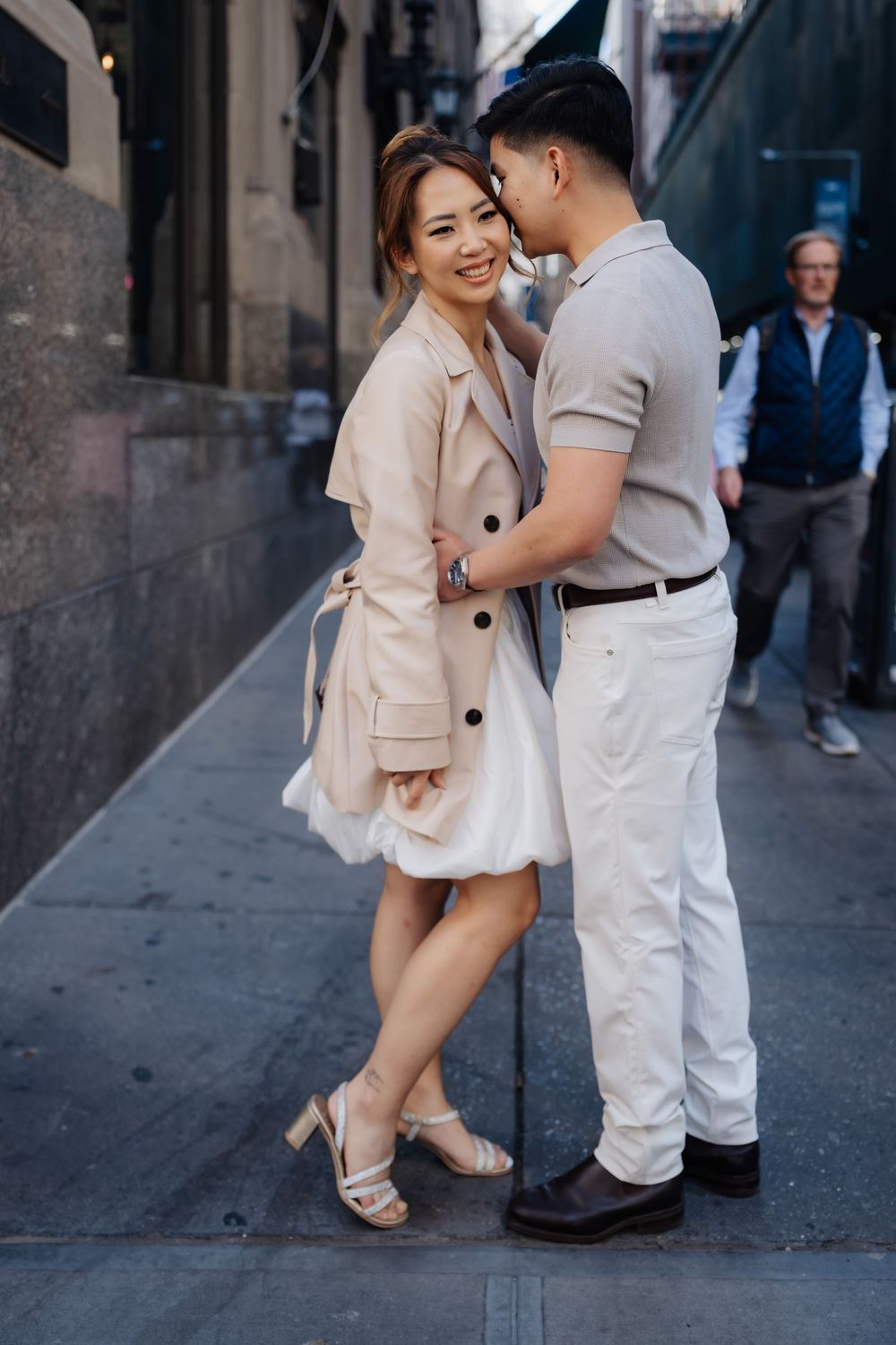 A smiling Asian couple embraces on a city sidewalk, woman in beige trench coat and white dress, man in gray polo and white pants.